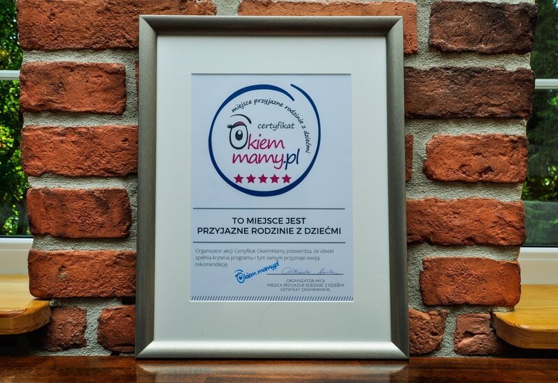 Srebrna ramka z certyfikatem „Okiem Mamy” na ceglanej ścianie przy oknie, potwierdzająca obiekt przyjazny rodzinom z dziećmi