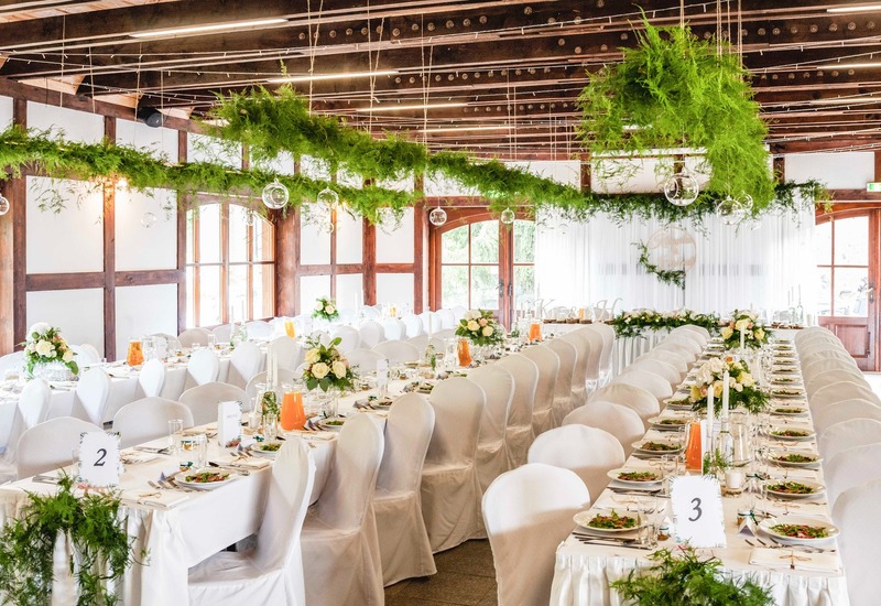Elegancka sala weselna z długimi białymi stołami, pokrowcami na krzesła, kwiatowymi aranżacjami, wiszącą zielenią i drewnianymi belkami.