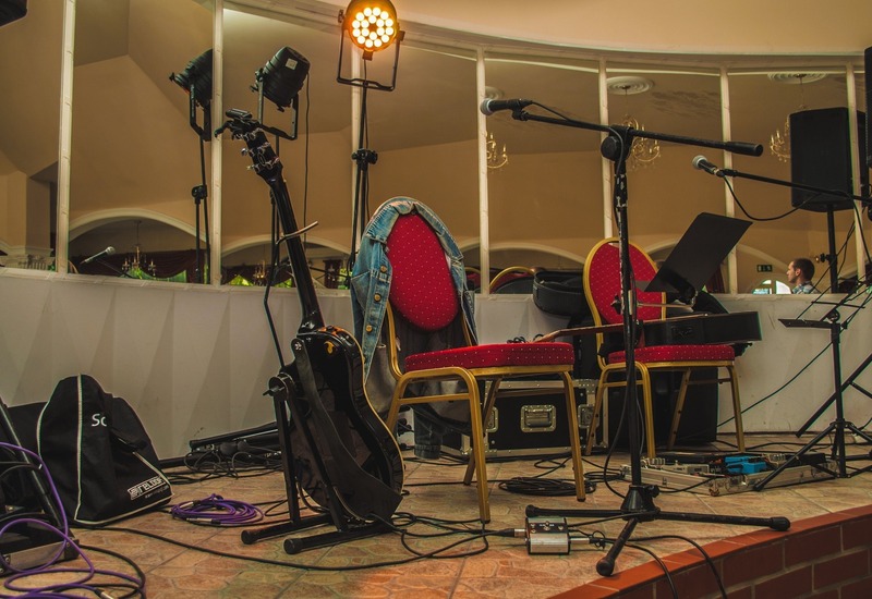 Scena hotelowa przygotowana na koncert: czerwone tapicerowane krzesła, gitary, mikrofony, oświetlenie i przewody.
