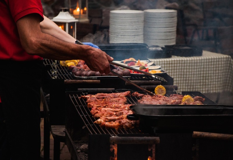 Bufetowy grill na zewnątrz — kucharz smaży marynowane mięsa i kukurydzę, tace z warzywami i stos talerzy na kraciastej obrusie.
