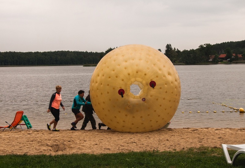 Piaszczysta plaża nad jeziorem z dużą żółtą nadmuchiwaną kulą (zorb) popychaną przez gości, leżaki i spokojne, zalesione brzegi.