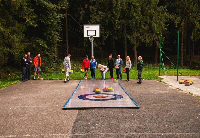 Grupa gra w plenerowy curling na boisku rekreacyjnym przy lesie, aktywność integracyjna pod koszem do koszykówki
