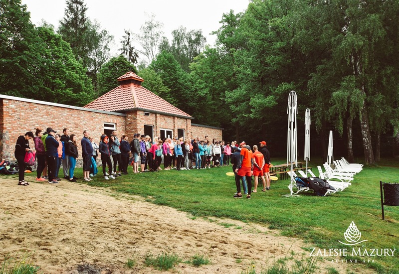 Goście ustawieni przy ceglanej pawilonie z czerwonym dachem, ratownicy w pomarańczowych strojach, leżaki i złożone parasole na trawiastej plaży