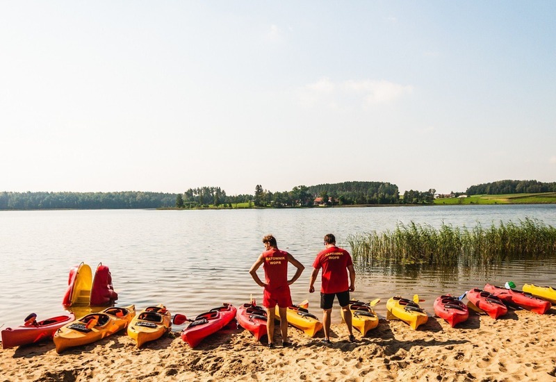 Spokojna plaża nad jeziorem z kolorowymi kajakami do wypożyczenia i dwoma ratownikami w czerwonych koszulkach.