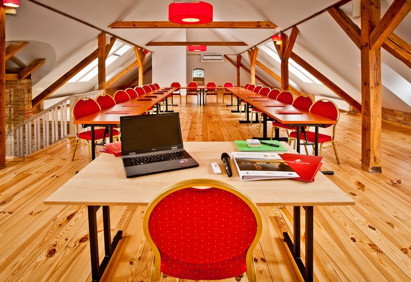 Sala konferencyjna na poddaszu z drewnianymi belkami, stołami i czerwonymi krzesłami, biurko z laptopem i mikrofonem.