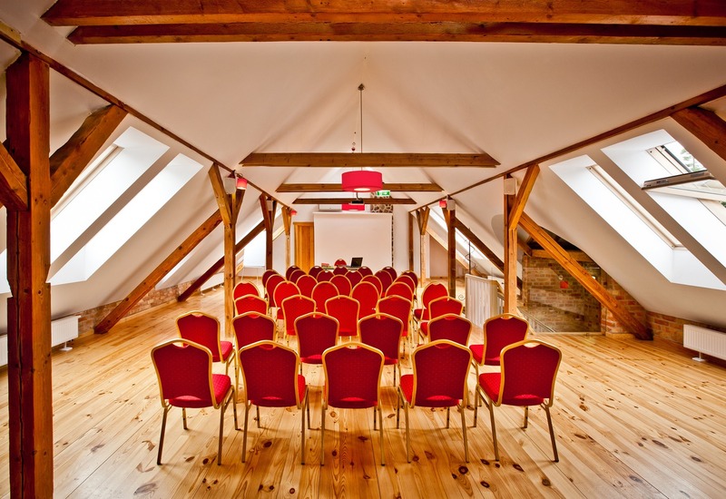 Sala konferencyjna na poddaszu z drewnianymi belkami, świetlikami, drewnianą podłogą i rzędami czerwonych krzeseł.