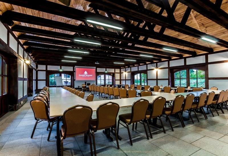 Przestronna rustykalna sala konferencyjna z układem podkowowym, licznymi krzesłami, odsłoniętymi belkami, ekranem i oknami z widokiem na ogród.