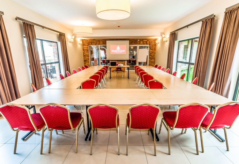 Sala konferencyjna w układzie U z czerwonymi krzesłami, projektorem, flipchartem, dużymi oknami, ceglaną ścianą i ciepłym oświetleniem