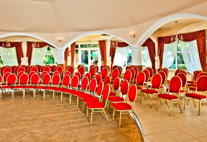 Elegancka sala bankietowa z rzędami czerwonych krzeseł, łukowymi kolumnami, żyrandolami i zasłonami z widokiem na ogród.