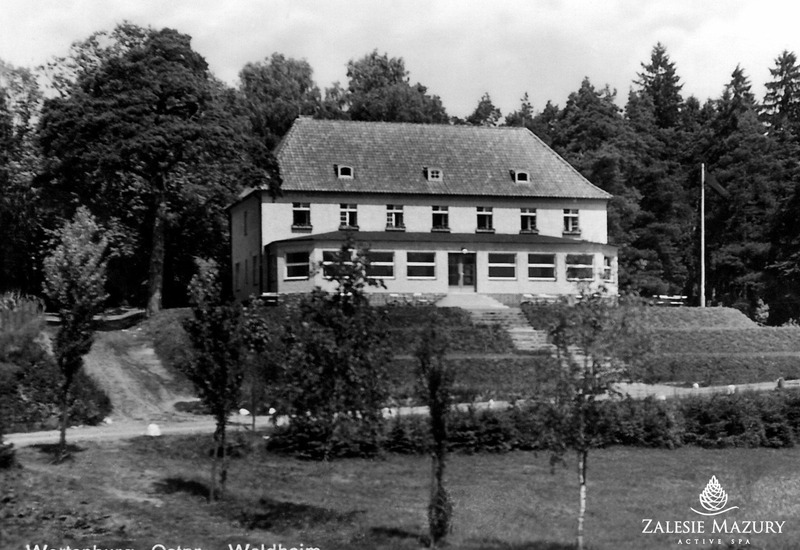 Historyczny, dwukondygnacyjny hotel z dachówką i dużymi oknami na parterze, pośród starych drzew i zadbanych trawników.