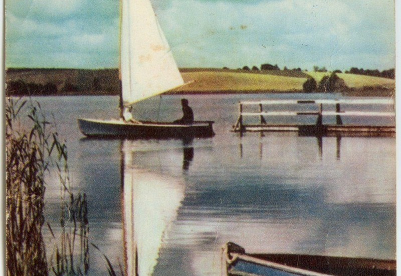 Żaglówka z jednym żeglarzem na spokojnym jeziorze przy drewnianym pomoście, mała łódź i trzciny na pierwszym planie, sielski widok idealny na relaks.