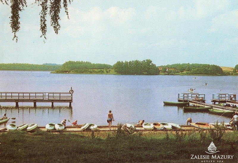 Pomost nad jeziorem z kajakami i łódkami; goście szykują sprzęt i odpoczywają przy spokojnej wodzie, w tle zalesione wyspy.