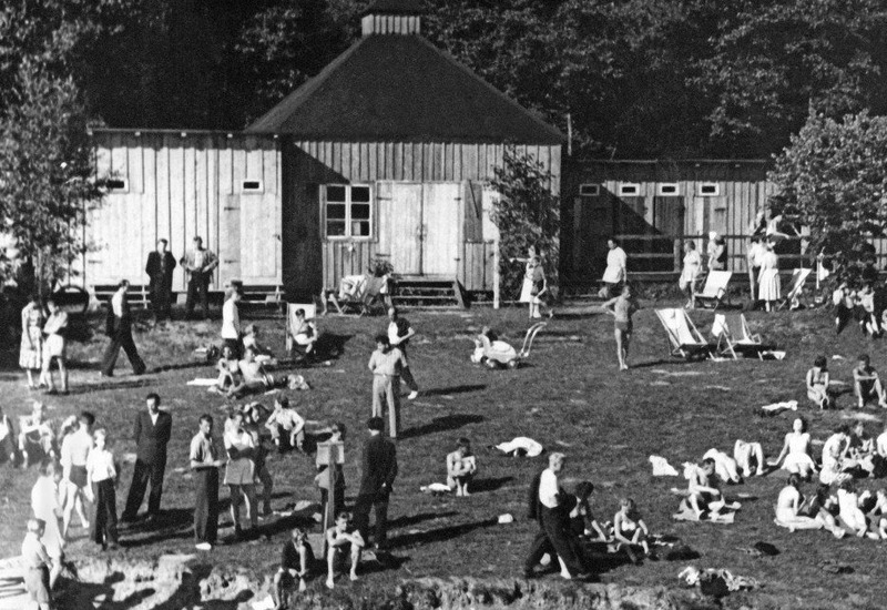 Plaża nad jeziorem z drewnianymi przebieralniami, leżakami oraz gośćmi opalającymi się i kąpiącymi; las w tle
