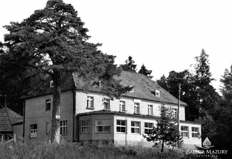 Zabytkowy budynek spa z dachówką, przeszklonym pawilonem i dużą sosną na tle lasu
