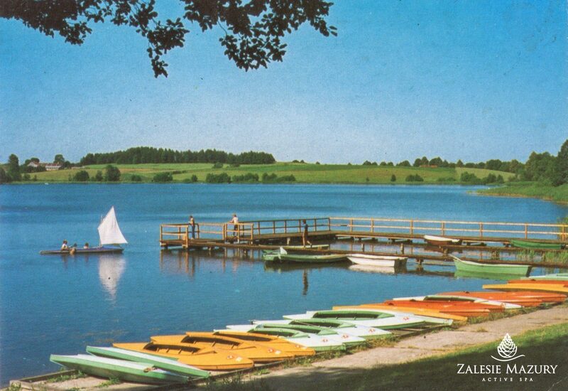 Pomost nad jeziorem z łodziami i kolorowymi kajakami na brzegu, goście spacerują, mały żagiel na spokojnej, błękitnej tafli.