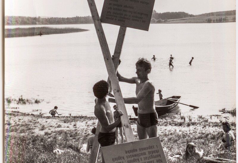 Plaża nad jeziorem, dzieci ustawiają tablicę, kąpiący się i łódka na spokojnej wodzie, trawiasty brzeg i odległy las