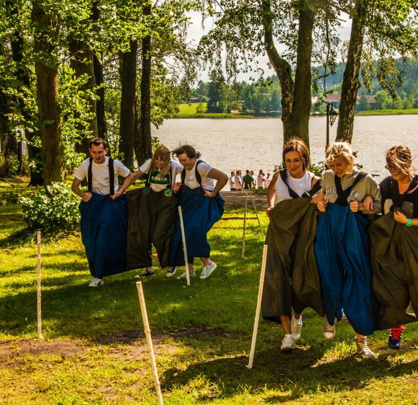 Grupa osób w wielkich workach ściga się po słonecznym trawniku nad jeziorem pod drzewami