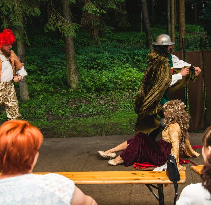 Plenerowy spektakl w lesie: aktorzy w strojach historycznych — aktor z czerwoną peruką, mężczyzna w hełmie, kobieta w aksamitnej sukni siedzi, widzowie na ławach.