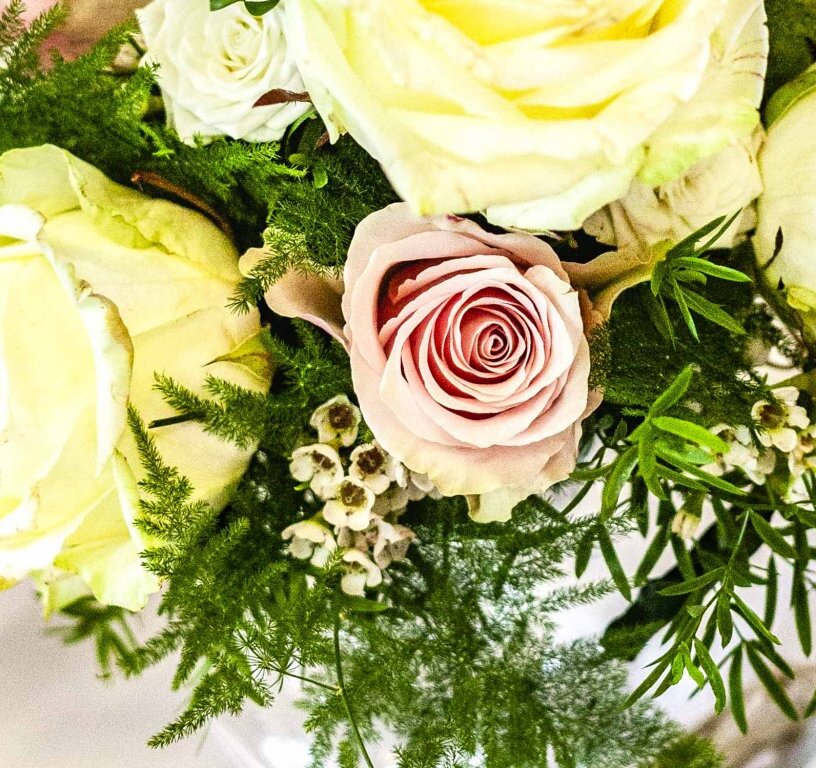 Elegant table centerpiece of pale yellow roses, a pink rose, green foliage and tiny white blooms on a white tablecloth.