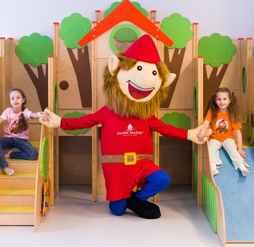 Hotel indoor kids' play area with wooden slides and a cheerful mascot holding hands with two children