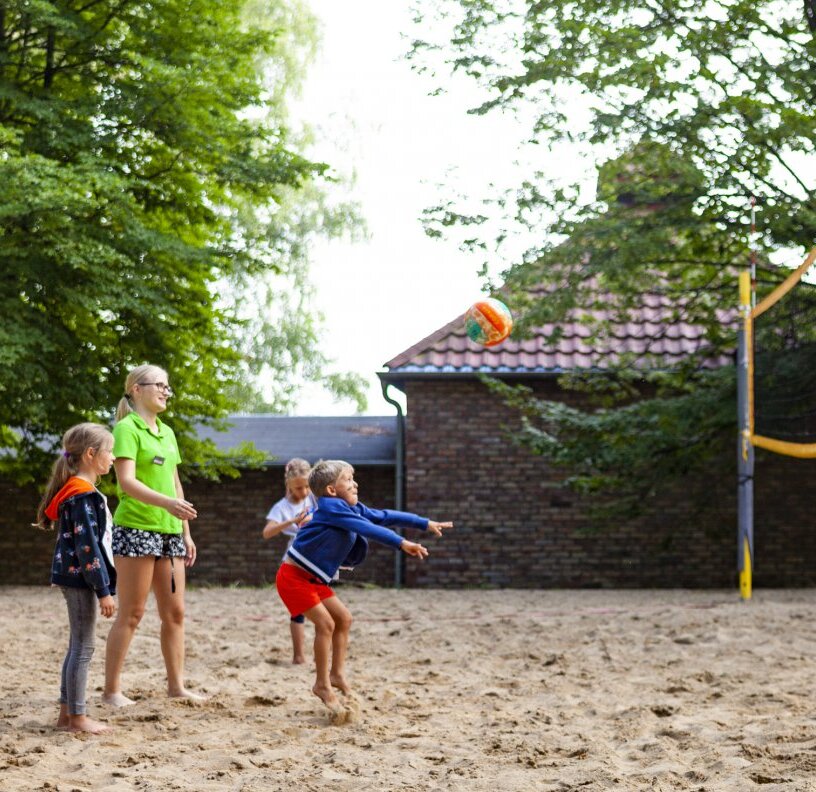 Children and a staff leader playing beach volleyball on a hotel sand court; instructor in green, trees and a tiled-roof building in background.