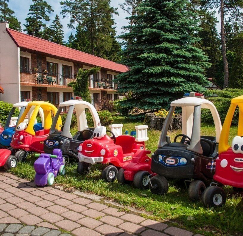 Row of colorful children's ride-on toy cars lined on lawn outside a family hotel with balcony rooms and pine-tree garden