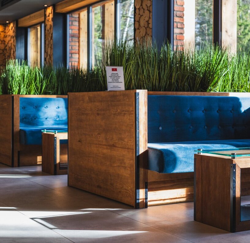 Hotel lounge booths with deep blue velvet seats, wooden dividers, glass tables and tall greenery by sunlit windows