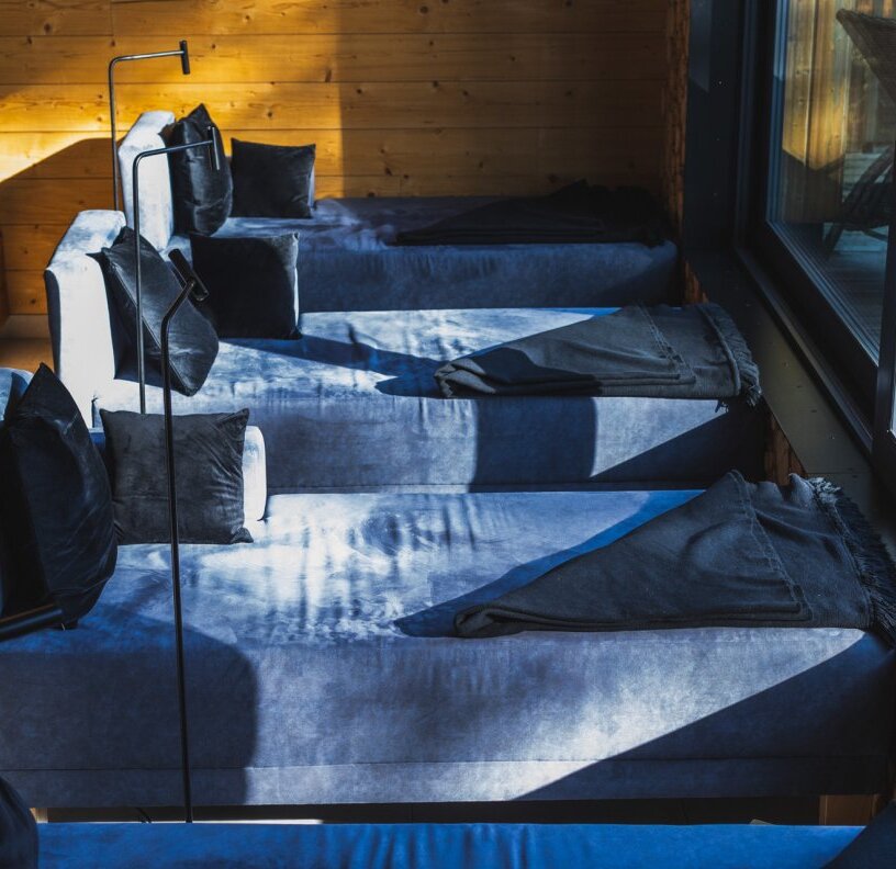Sunlit row of blue velvet daybeds with black throws and cushions in a wood-paneled relaxation lounge by a large window