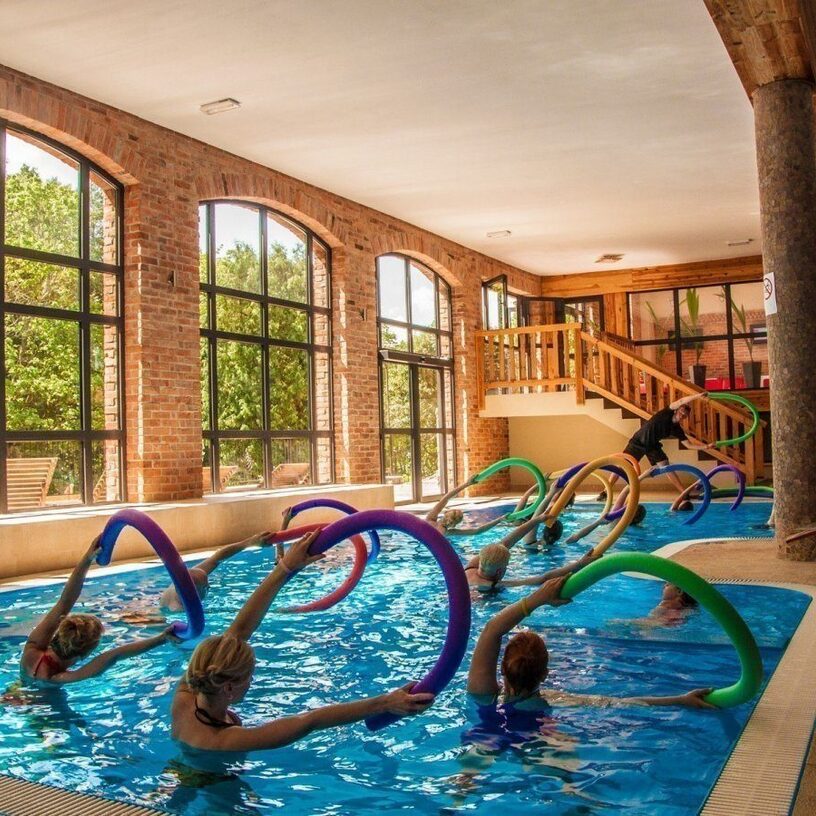 Indoor hotel pool with arched brick windows, group aqua fitness class using colorful pool noodles led by instructor.