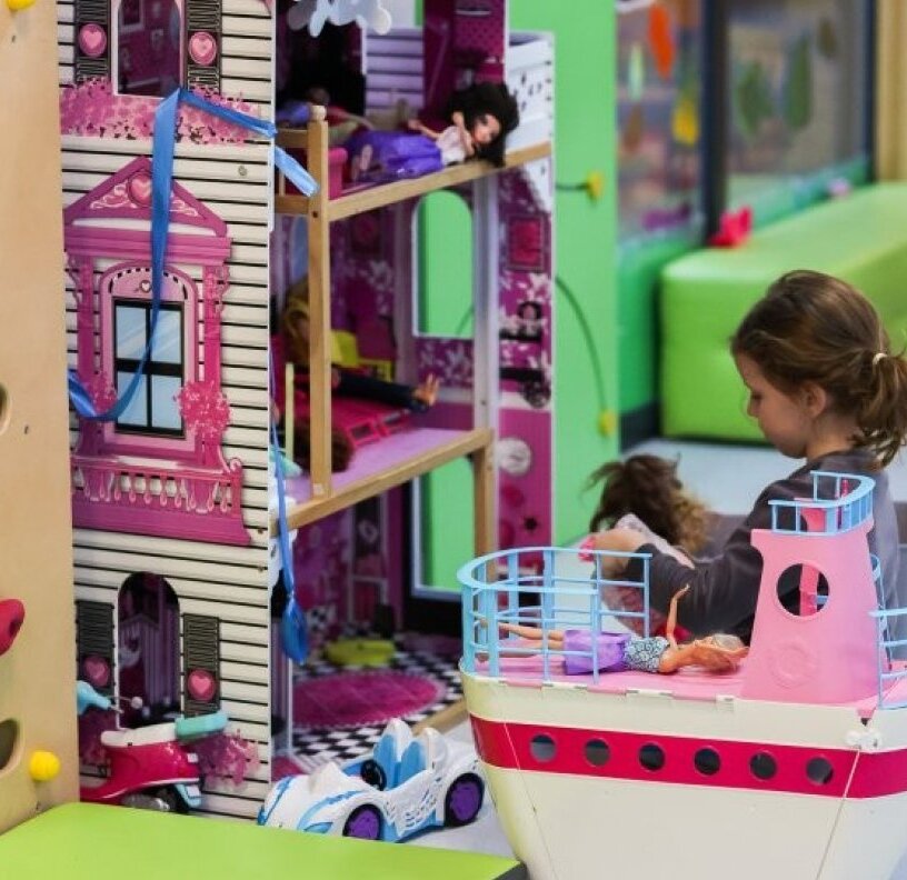 Colorful children's playroom with dollhouse, toy boat and climbing wall; child playing with dolls on padded bench