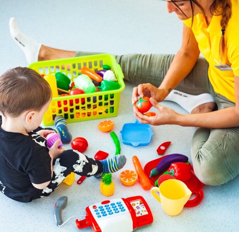 Toddler and staff member in hotel kids' club play area, sitting on the floor with a basket of colorful plastic fruits and toys during supervised playtime
