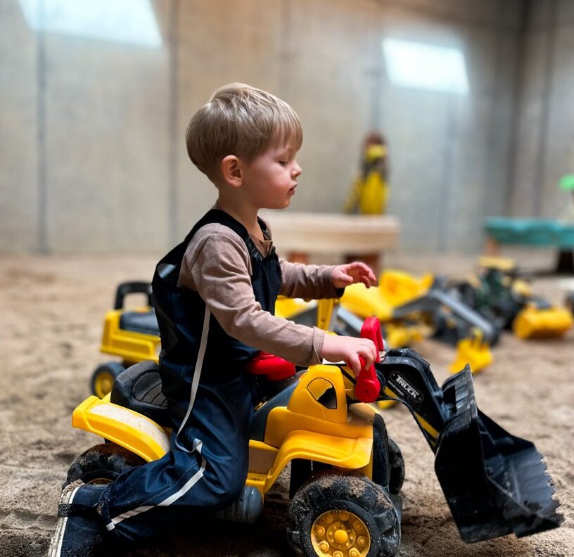 Toddler playing on a yellow ride-on excavator in the hotel's indoor sand play area surrounded by toy construction trucks