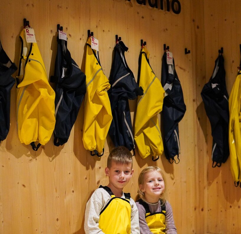 Children in yellow rental waterproof suits smiling in a wooden cloakroom with rows of yellow and navy jackets hanging.