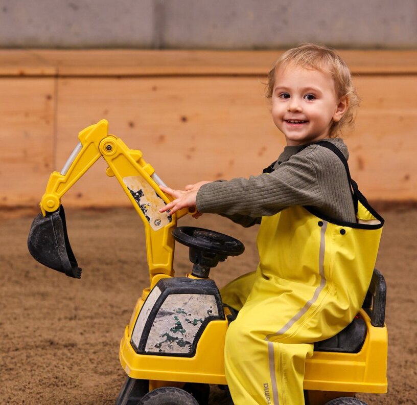 Smiling toddler in yellow rain suit driving a toy excavator in a hotel's indoor kids' play area with soft sand floor.