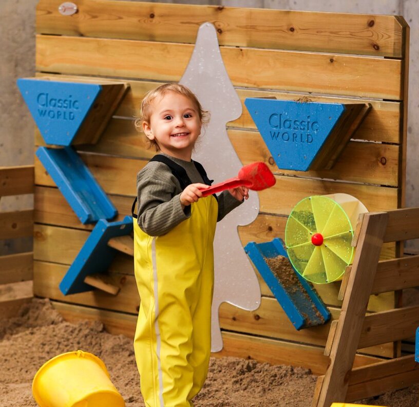 Smiling toddler in yellow rain suit playing with a red shovel in indoor sand play area with interactive wall toys and bucket