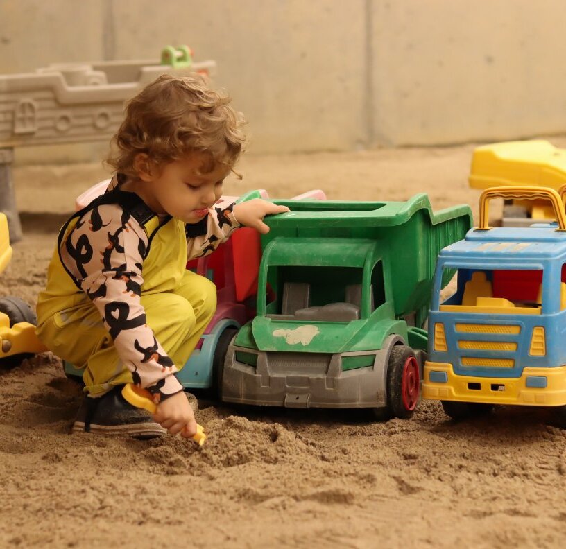 Toddler playing with plastic dump trucks in a sandy indoor kids' play area at a family-friendly hotel.