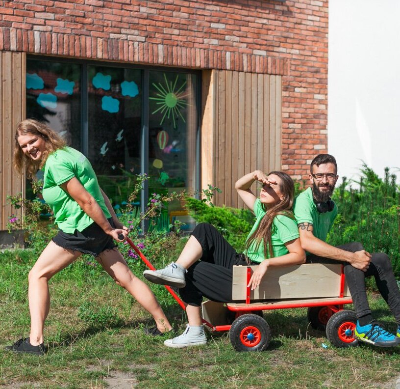 Three people in green shirts playing outdoors: one woman pulls a red wagon while two others sit inside near a brick building with playful window art.