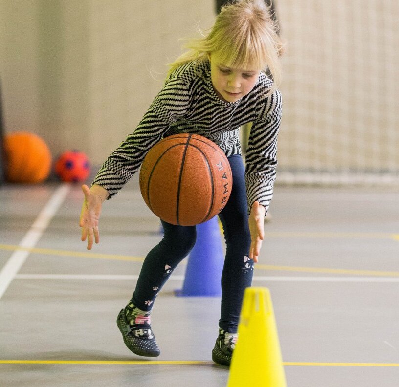 Young girl dribbling a basketball through training cones in an indoor gym, focused on the ball