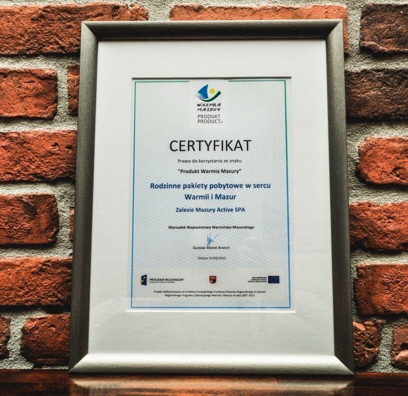 Oprawiony certyfikat Zalesie Mazury Active SPA za rodzinne pakiety pobytowe, zawieszony na surowej ceglanej ścianie