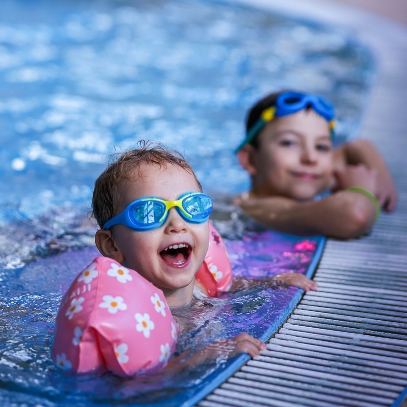 Two children in a shallow indoor pool, one laughing at the edge wearing pink floral arm floaties and blue swim goggles