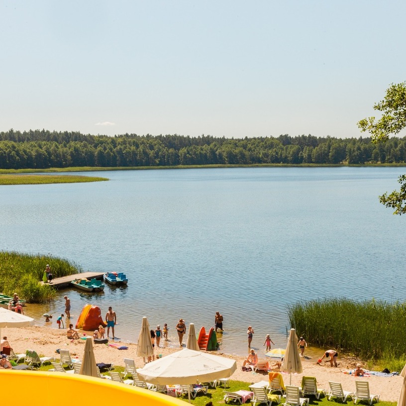 Sandy hotel beach on a calm lake with sun loungers and umbrellas, families swimming, paddleboats and a small dock framed by reeds and forest.