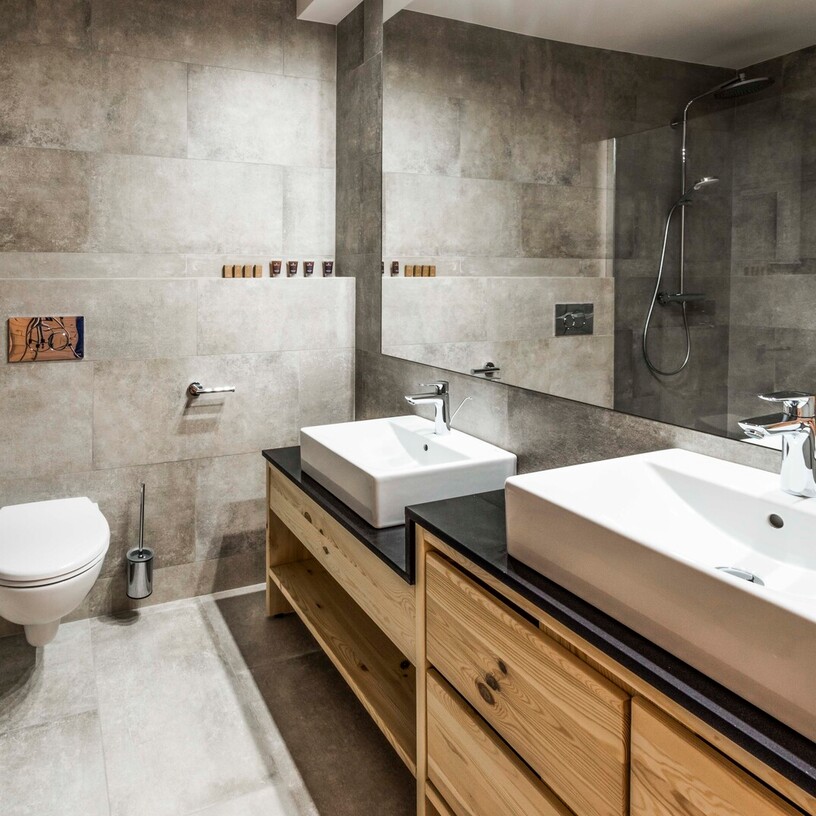 Modern hotel bathroom with double rectangular sinks on wooden vanity, large mirror, walk-in rain shower, wall-mounted toilet and toiletries.