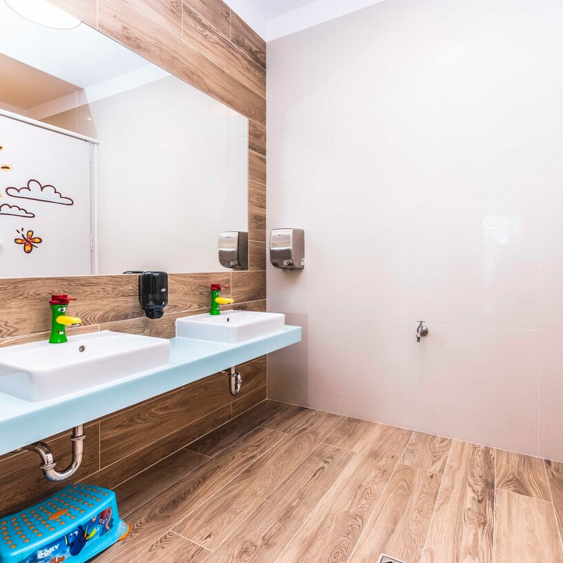 Bright family bathroom with twin child-height sinks, colorful playful faucets, large mirror, wood-look tiles and kids' step stool.