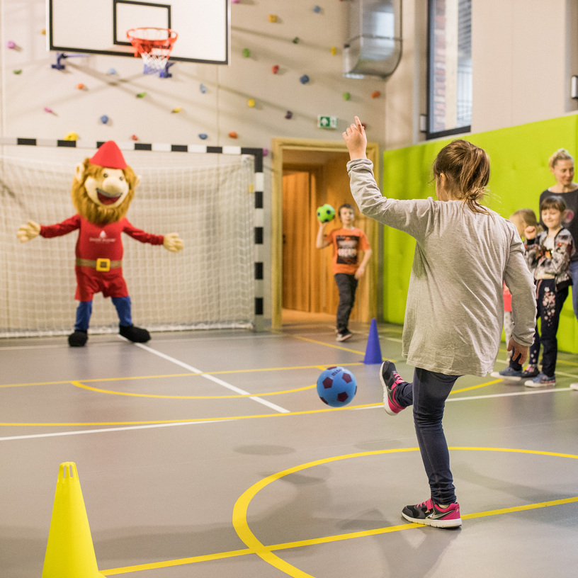 Hotel kids' club sports hall with family watching as a child kicks a ball toward a smiling lion mascot in the goal.