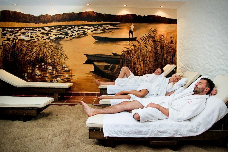 Hotel spa relaxation room with sand floor, cushioned loungers and three guests in white robes resting under a warm lakeside mural.