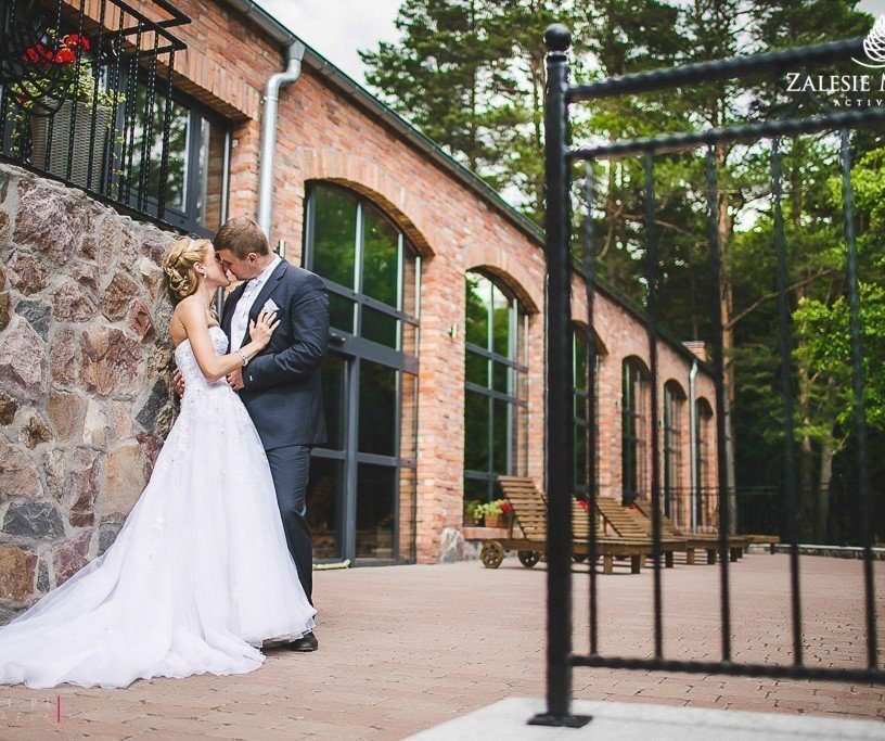Bride and groom kissing on a spa resort terrace by a brick building with arched windows, lounge chairs and pine-forest backdrop
