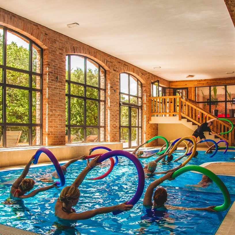 Kryty basen hotelowy z ceglanymi łukowymi oknami i antresolą, zajęcia aqua aerobiku z kolorowymi makaronami do pływania