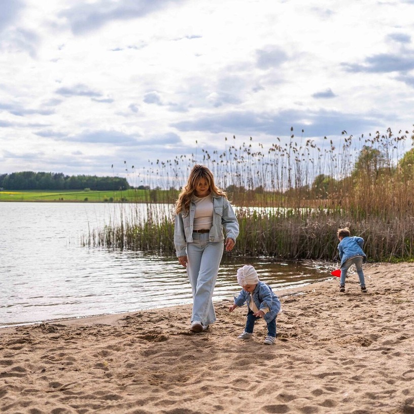 Piaszczysta plaża nad jeziorem z trzcinami i leżakiem; matka spaceruje, dwoje dzieci bawi się przy brzegu, widok na wiejską zieleń.