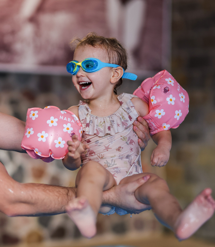 Laughing toddler in blue goggles and pink floral arm-floats held above a hotel's indoor pool, playful family swim.
