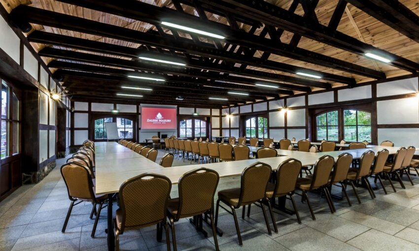 Rustic conference room with exposed wooden beams, U-shaped tables, padded chairs, projector screen and flipchart, garden views